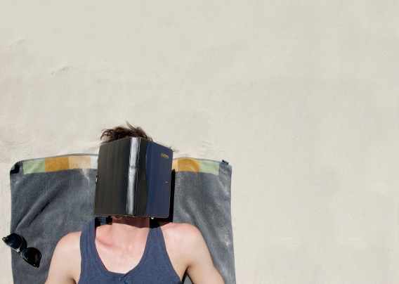 Man Lying Down On A Towel At The Beach With Book Covering Face